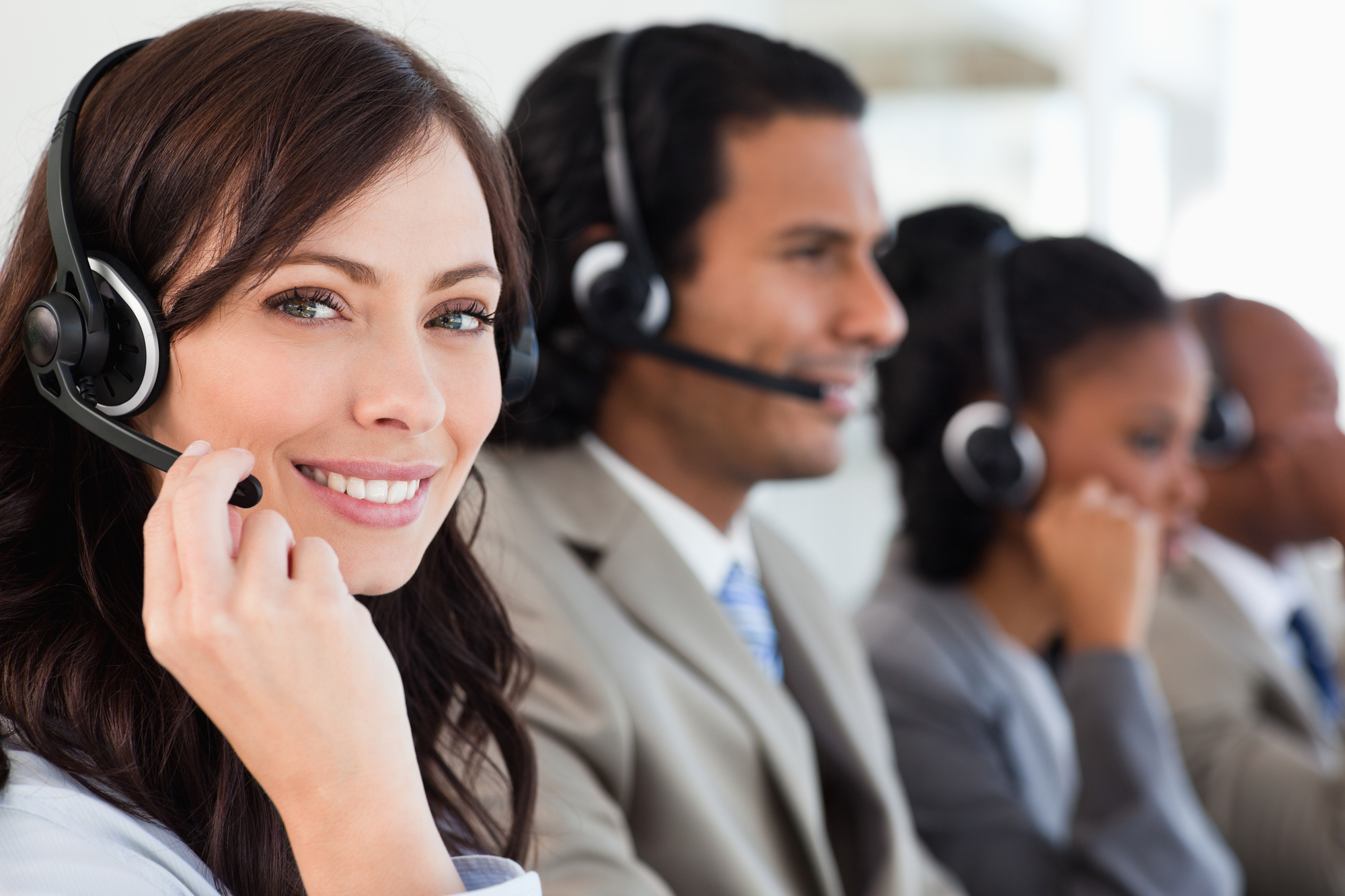 A row of workers sit on the phone and take phone calls while wearing their headsets. The woman on the far left is in focus and smiling at the camera.